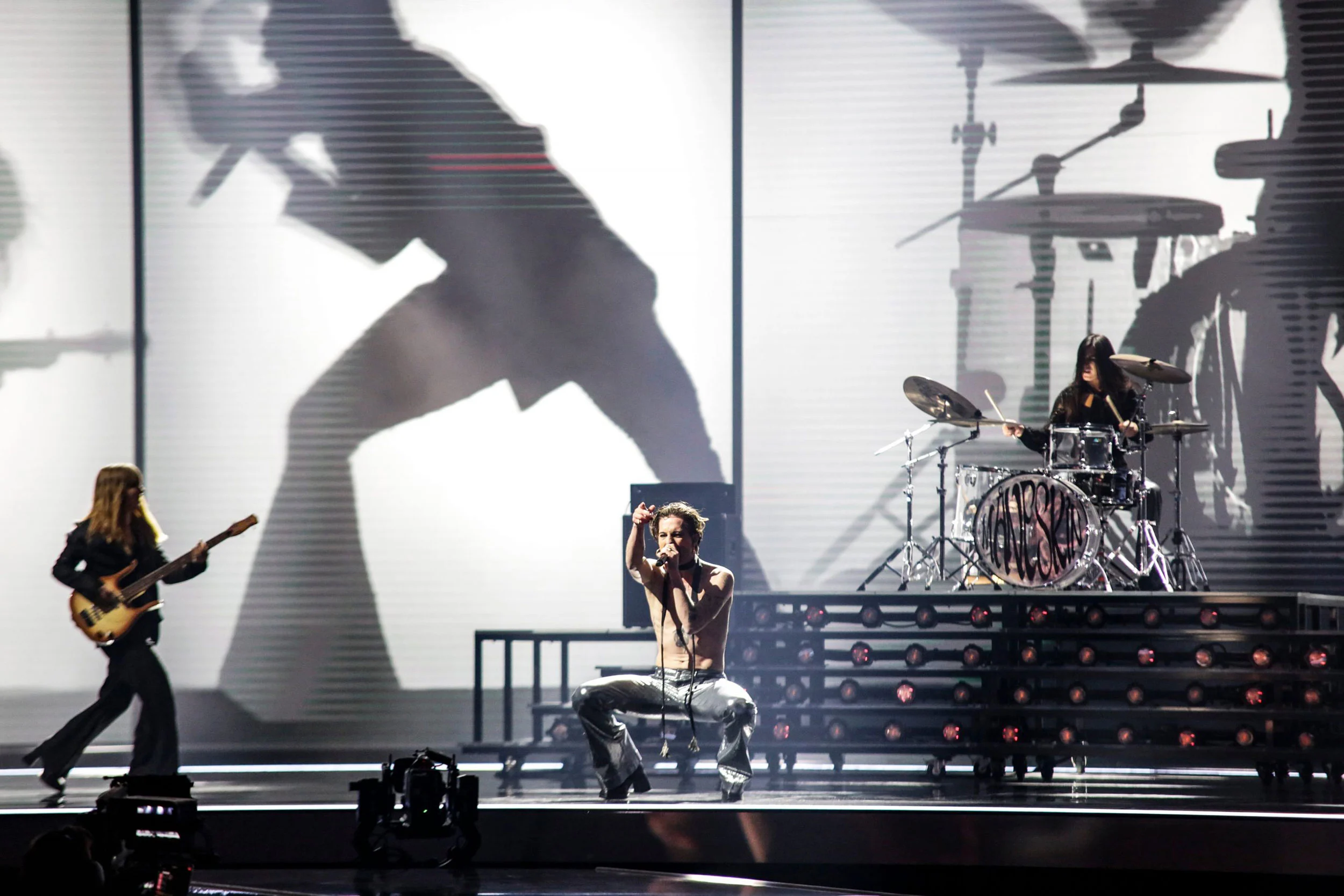 Cantante accovacciato in proscenio, chitarrista e batterista dietro, sagome della band proiettate sul fondale – Måneskin all'Eurovision 2021, Rotterdam, Paesi Bassi – scenografia per concerti