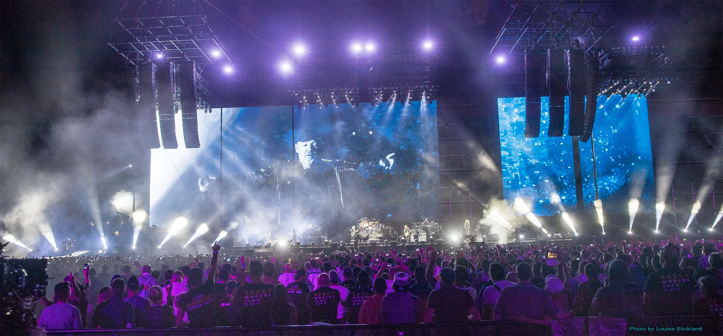 Cantante ripreso sullo schermo principale e su uno schermo laterale virato in blu, riflettori bianchi che spazzano il pubblico – Vasco Rossi Modena Park, Italia, 2017 – scenografia per concerti