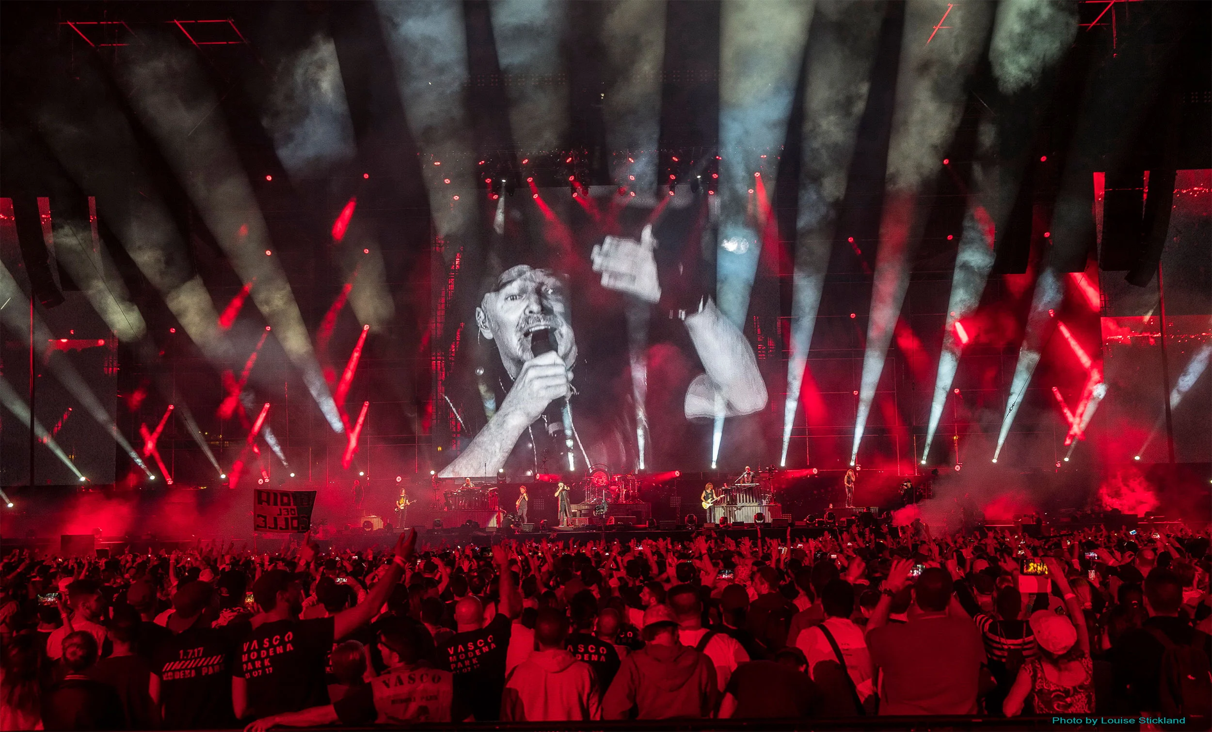 Vasco Rossi su uno schermo gigante sopra la band, palco immerso in luce rossa e fumo durante il concerto – Vasco Rossi Modena Park, Italia, 2017 – scenografia per concerti
