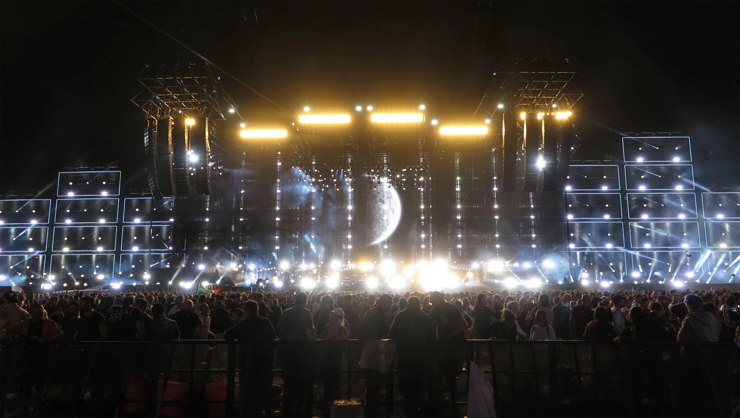 Vista notturna dell'intero palco con barre di luce dorate, torri laterali rivestite di schermi e l'immagine di una luna al centro – Vasco Rossi Modena Park, Italia, 2017 – scenografia per concerti