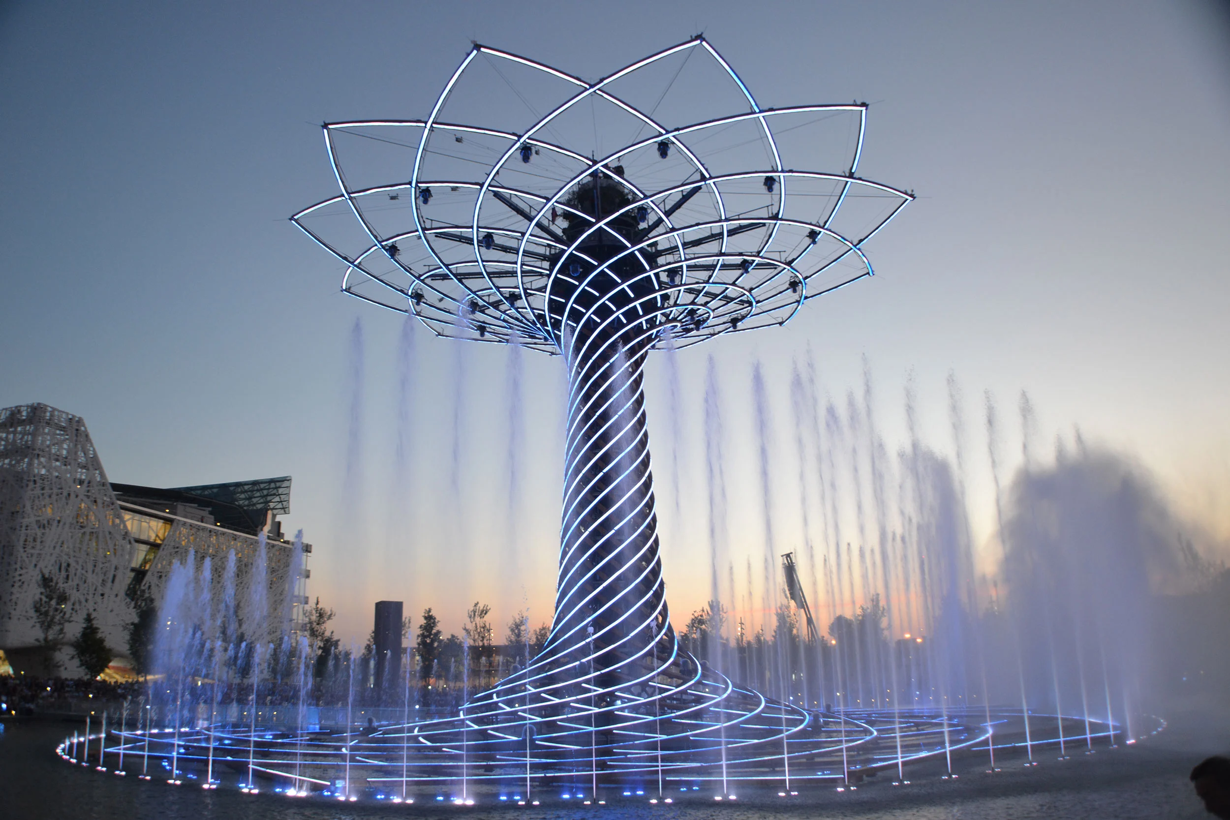 Albero della Vita al crepuscolo: chioma a reticolo d'acciaio e tronco a spirale sopra un anello di getti d'acqua – Albero della Vita all'Expo 2015, Milano, Italia – progettazione di installazioni
