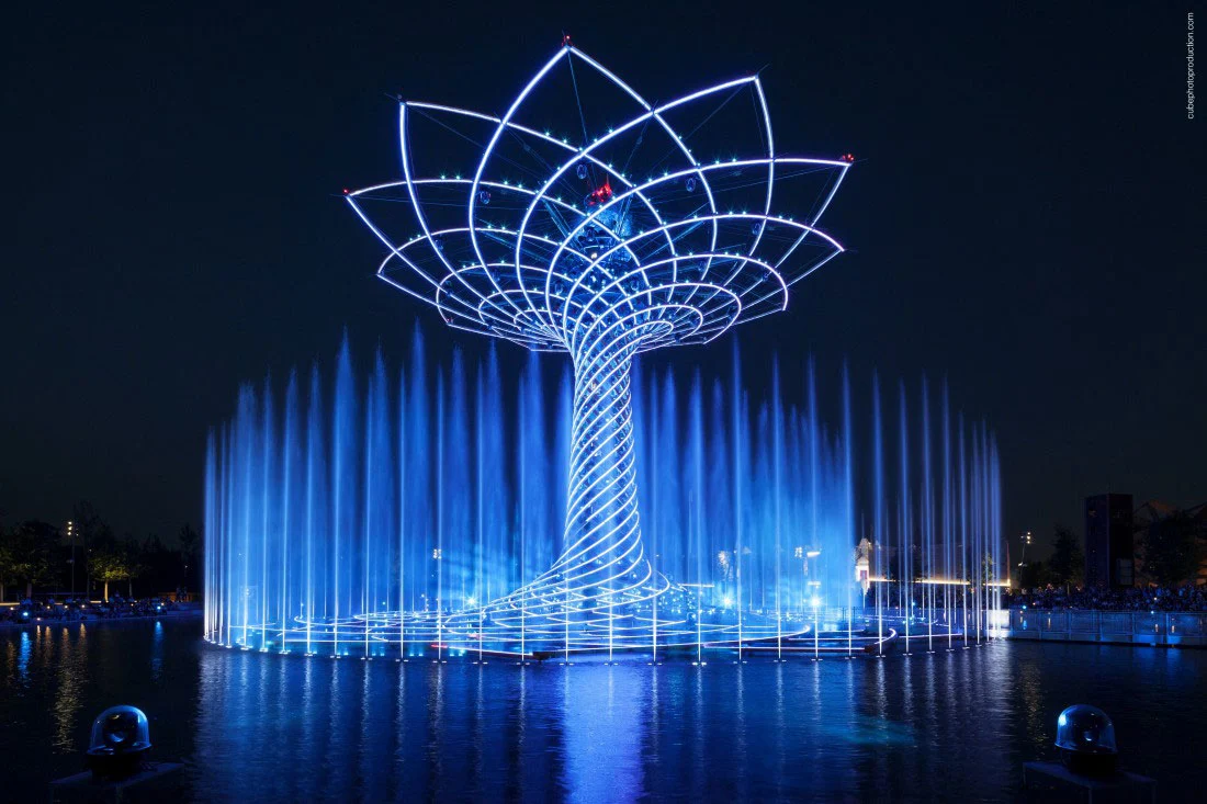 Albero della Vita illuminato di blu di notte, getti della fontana intorno alla base riflessi nel lago – Albero della Vita all'Expo 2015, Milano, Italia – progettazione di installazioni