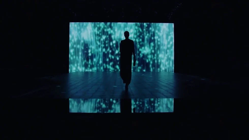 Sagoma di una modella che cammina verso una parete LED con scie di luce verde acqua, riflessa sul pavimento – Giorgio Armani SS21 Donna, Milano, Italia, 2021 – scenografia per sfilate