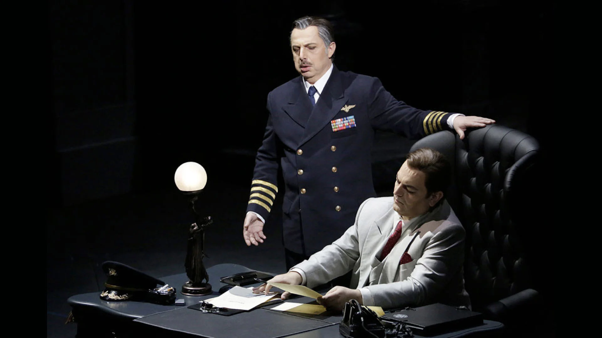 Ufficiale in uniforme blu navy in piedi accanto a un uomo seduto in abito chiaro a una scrivania con una lampada a globo – Un Ballo in Maschera al Teatro Bolshoi, Mosca, Russia, 2018 – scenografia d'opera