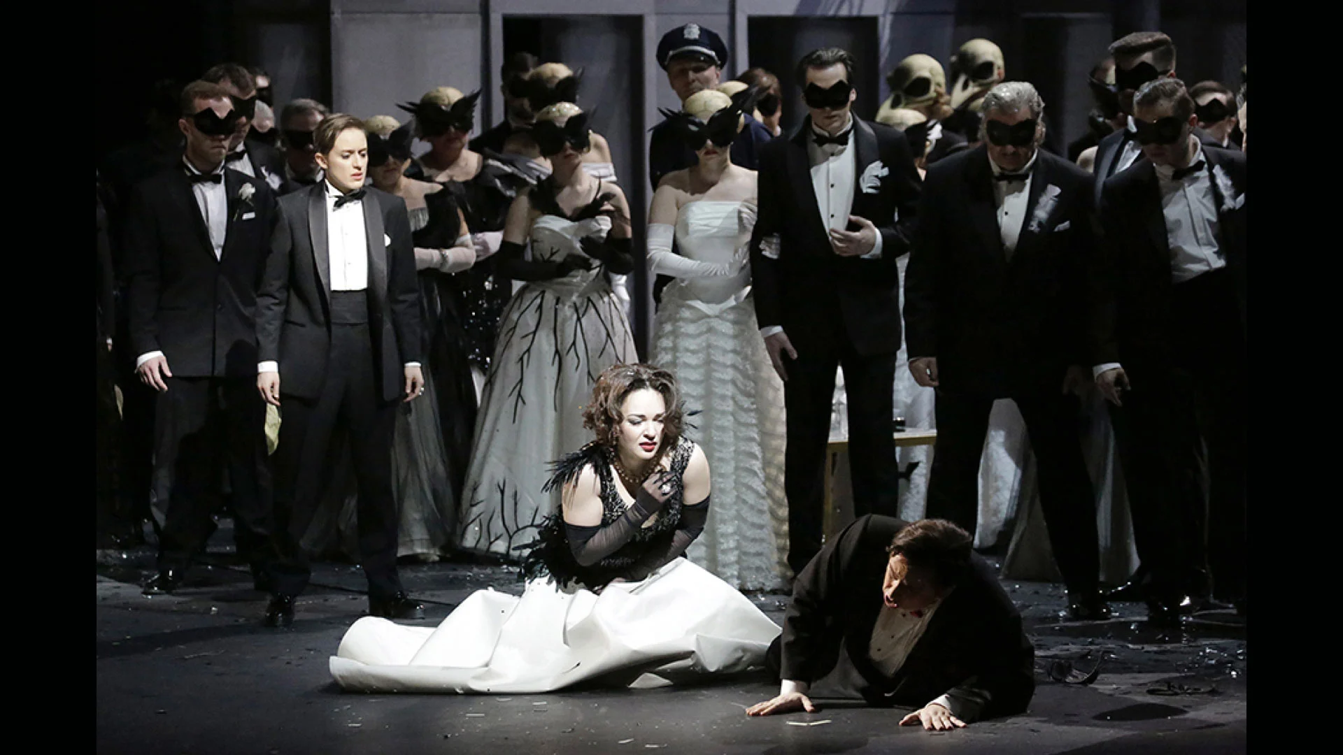 Donna in abito bianco inginocchiata accanto a un uomo a terra, circondata da invitati in maschera in abito da sera – Un Ballo in Maschera al Teatro Bolshoi, Mosca, Russia, 2018 – scenografia d'opera