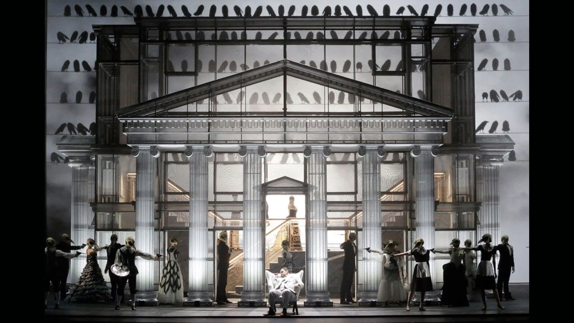 Scenografia con facciata di tempio neoclassico e sagome di uccelli sulle pareti, invitati in maschera schierati ai due lati – Un Ballo in Maschera al Teatro Bolshoi, Mosca, Russia, 2018 – scenografia d'opera