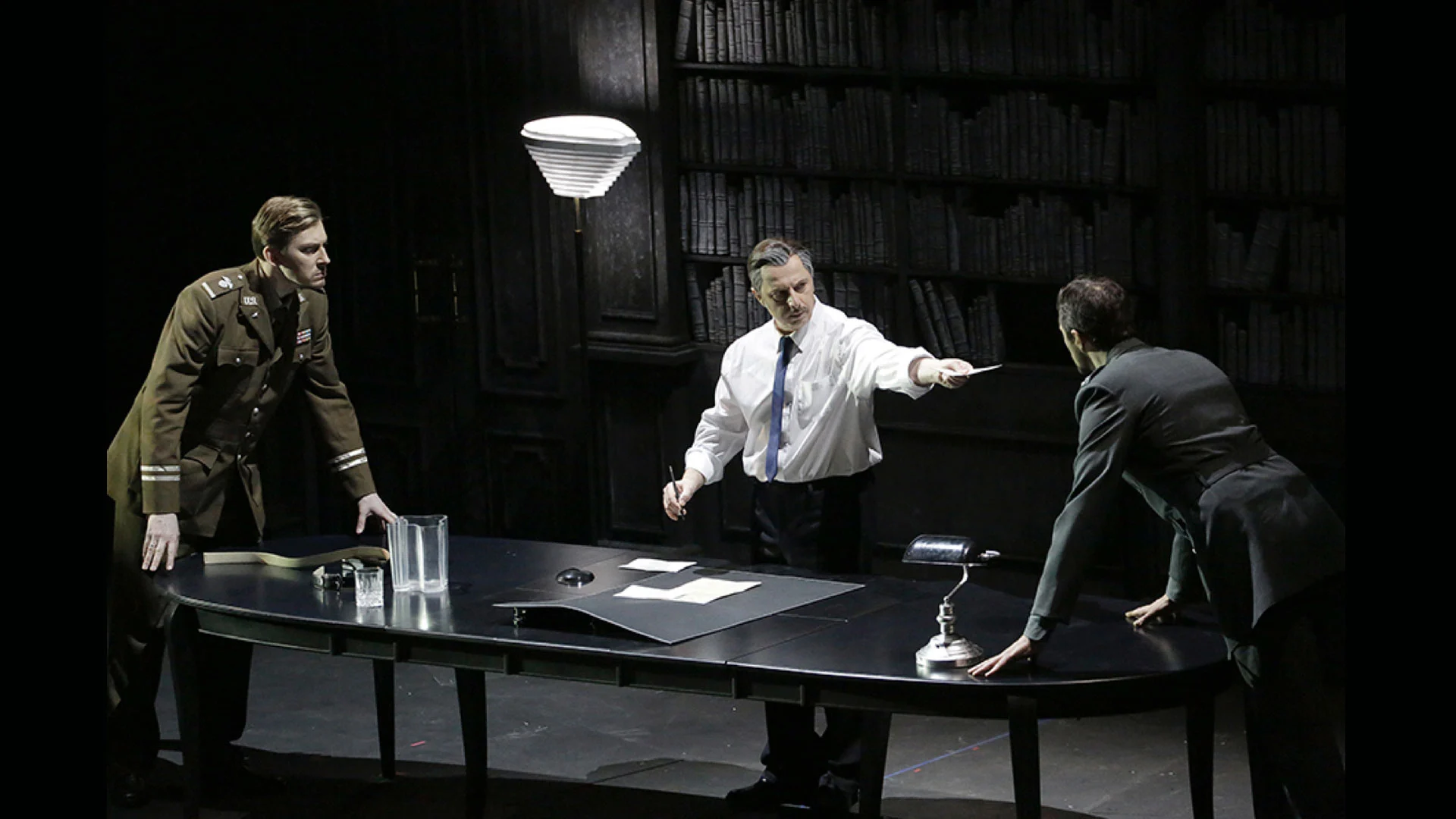 Tre artisti intorno a un tavolo ovale scuro in uno studio rivestito di librerie, illuminato da una lampada a stelo – Un Ballo in Maschera al Teatro Bolshoi, Mosca, Russia, 2018 – scenografia d'opera