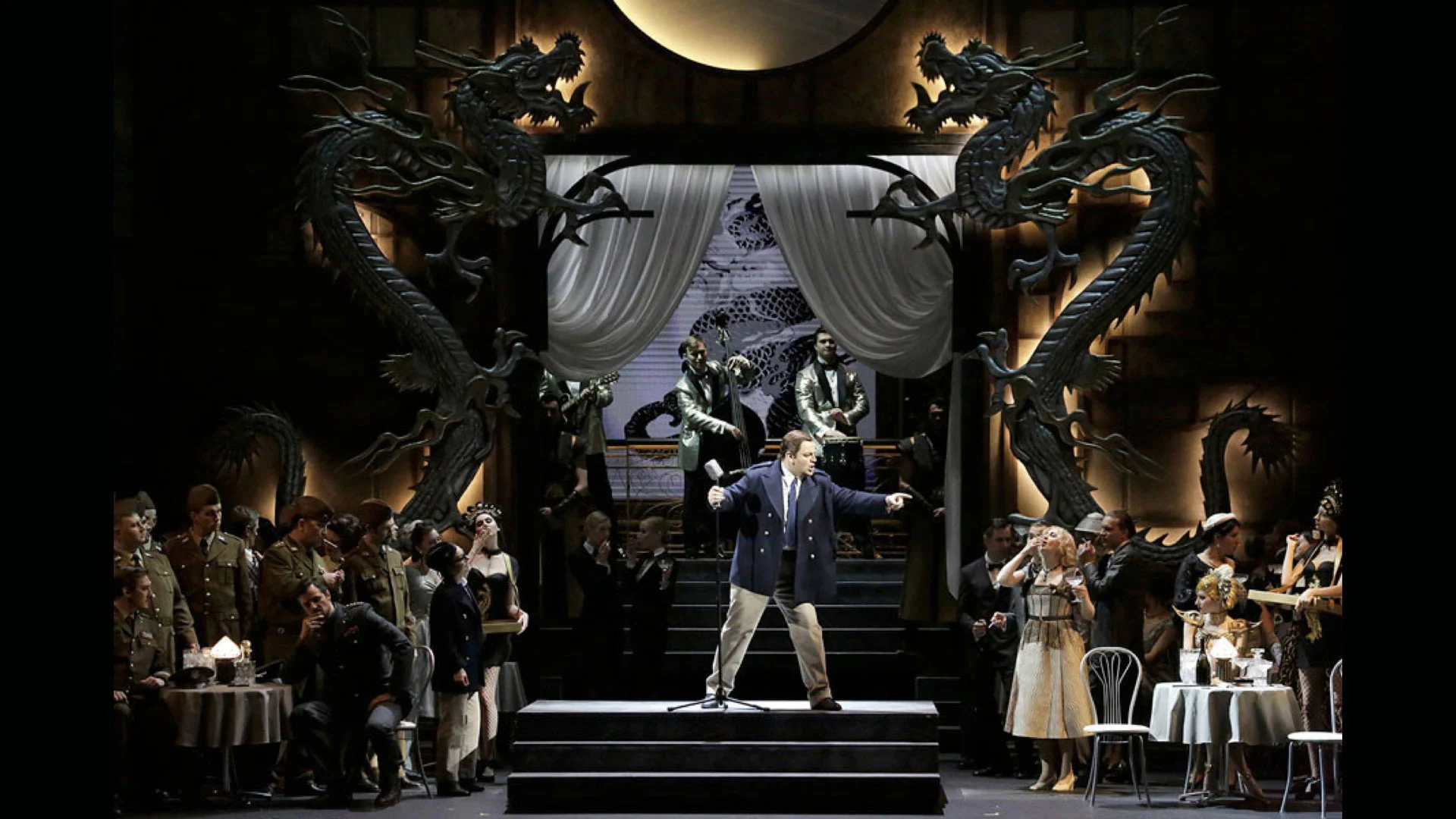 Scenografia con due gigantesche sculture di draghi ai lati di una porta drappeggiata, soldati e ospiti ai tavolini di un caffè – Un Ballo in Maschera al Teatro Bolshoi, Mosca, Russia, 2018 – scenografia d'opera