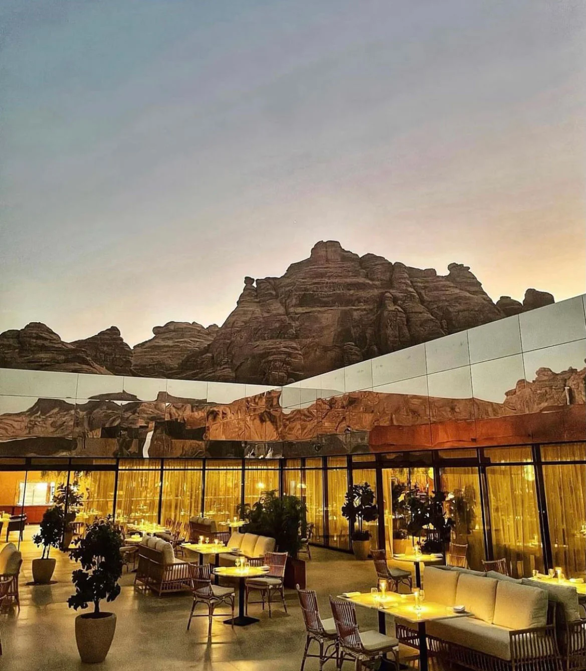 Terrazza del ristorante al crepuscolo con tavoli illuminati e sedie in rattan, falesie riflesse nella parete a specchio sopra – Maraya Concert Hall, AlUla, Arabia Saudita, 2019 – architettura