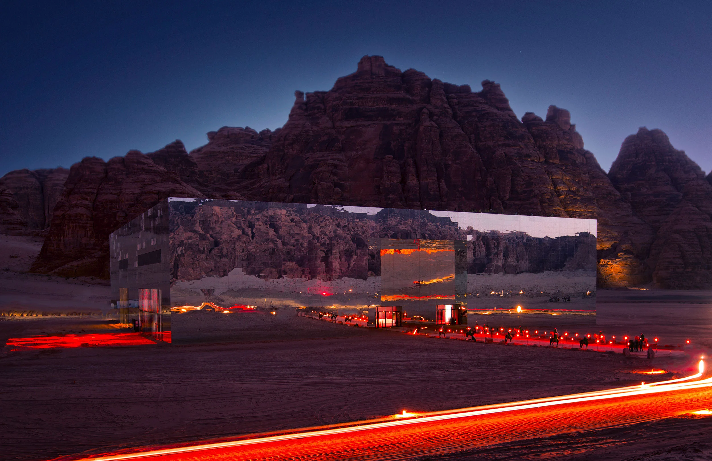 Maraya di notte: edificio a specchio che riflette le falesie, con scie di luce rossa lungo la strada nel deserto – Maraya Concert Hall, AlUla, Arabia Saudita, 2019 – architettura