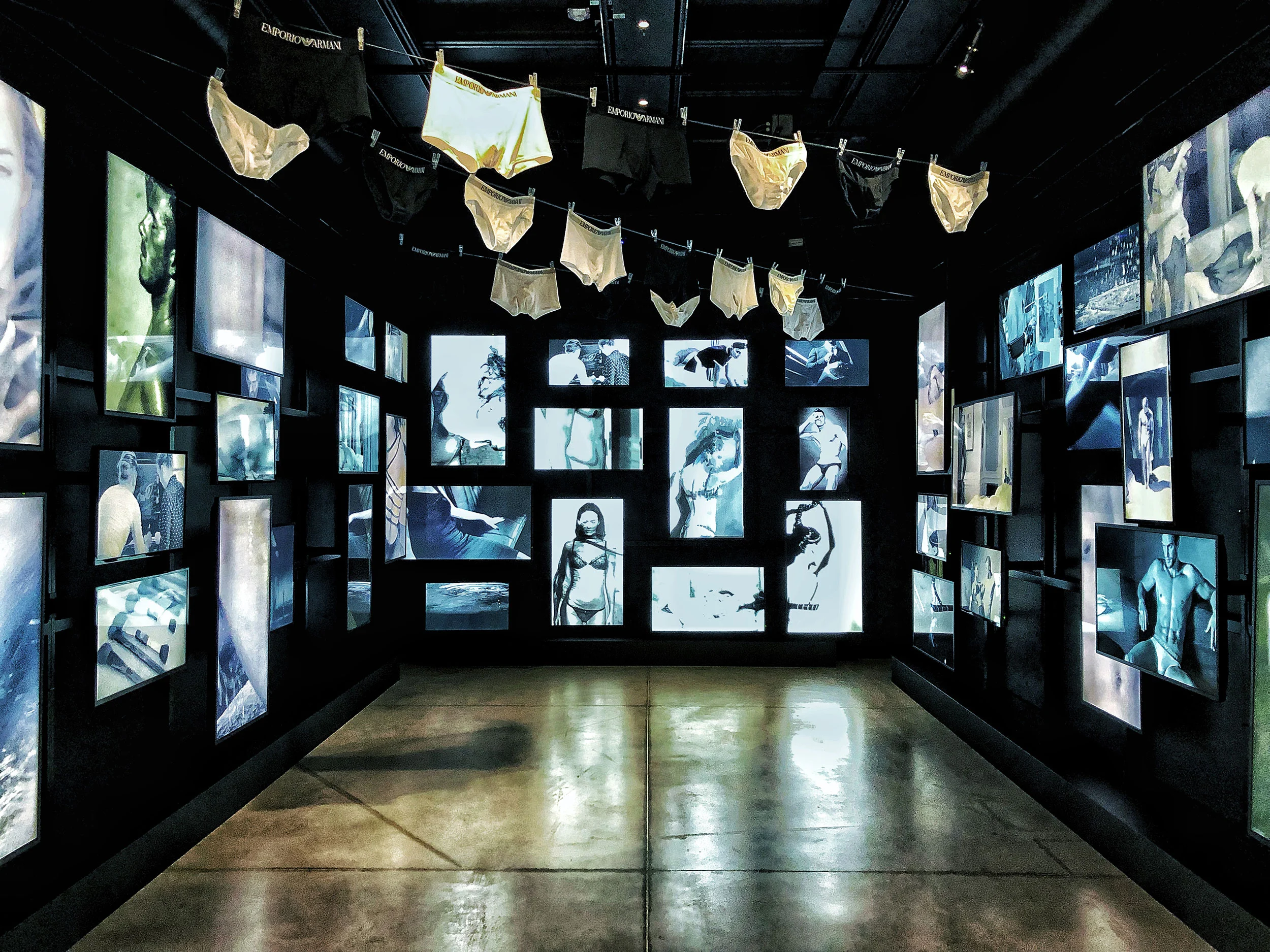 Sala buia di immagini pubblicitarie di intimo retroilluminate, con slip stesi su un filo in alto – Emporio Armani The Way We Are, Milano, Italia, 2020 – allestimento espositivo