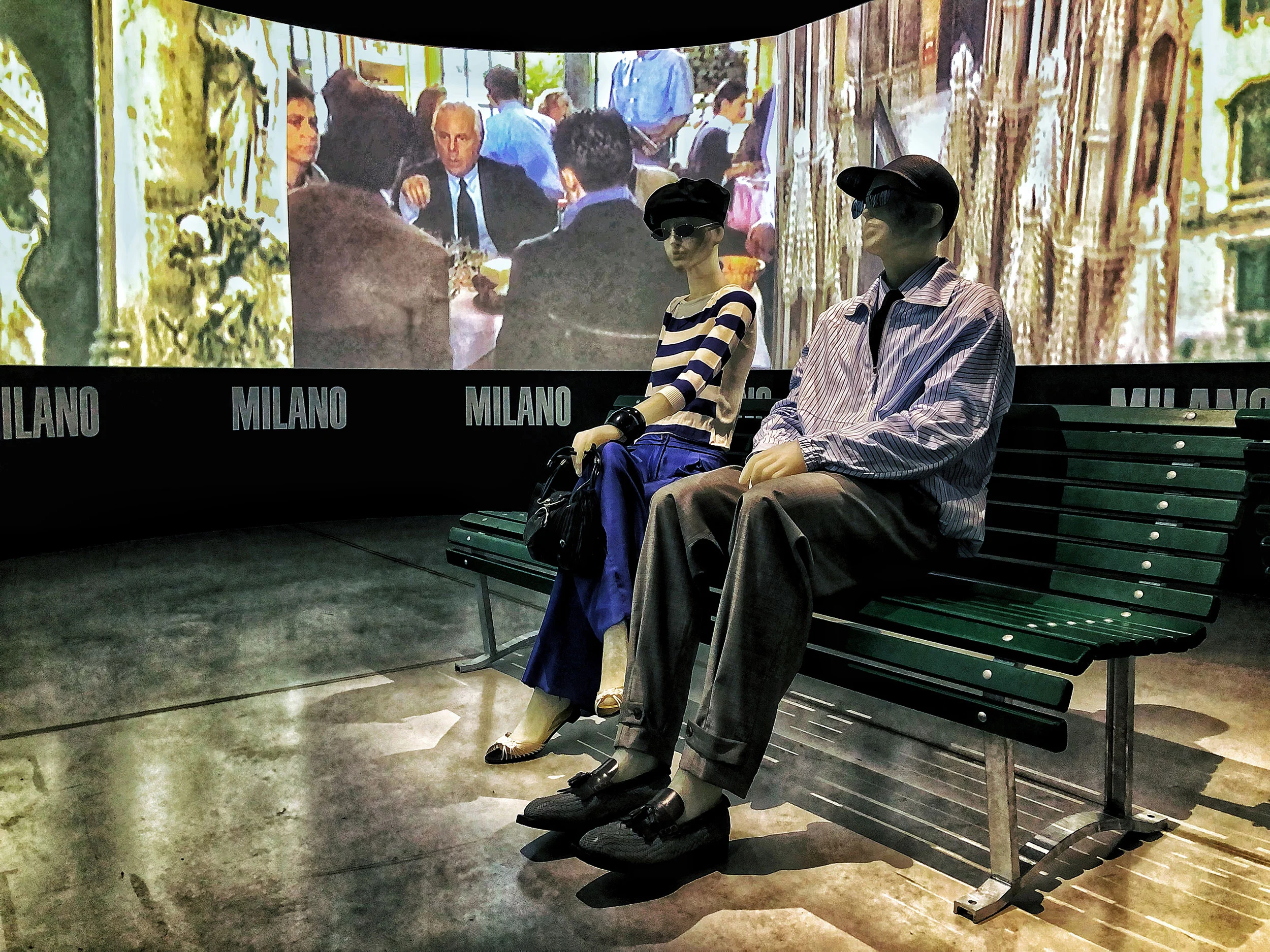 Due manichini su una panchina verde da parco davanti a uno schermo curvo con scene di Milano e la scritta Milano – Emporio Armani The Way We Are, Milano, Italia, 2020 – allestimento espositivo