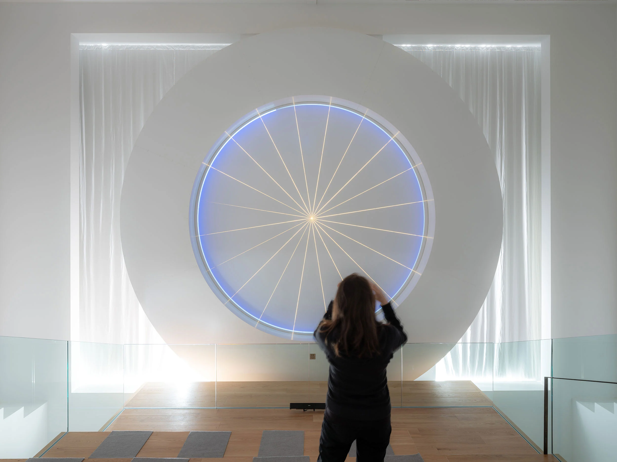 Visitatore che fotografa un'apertura circolare illuminata di blu con linee a raggiera, in una parete bianca tra due tende – Lumen Museo della fotografia di montagna, Plan de Corones, Italia, 2018 – architettura