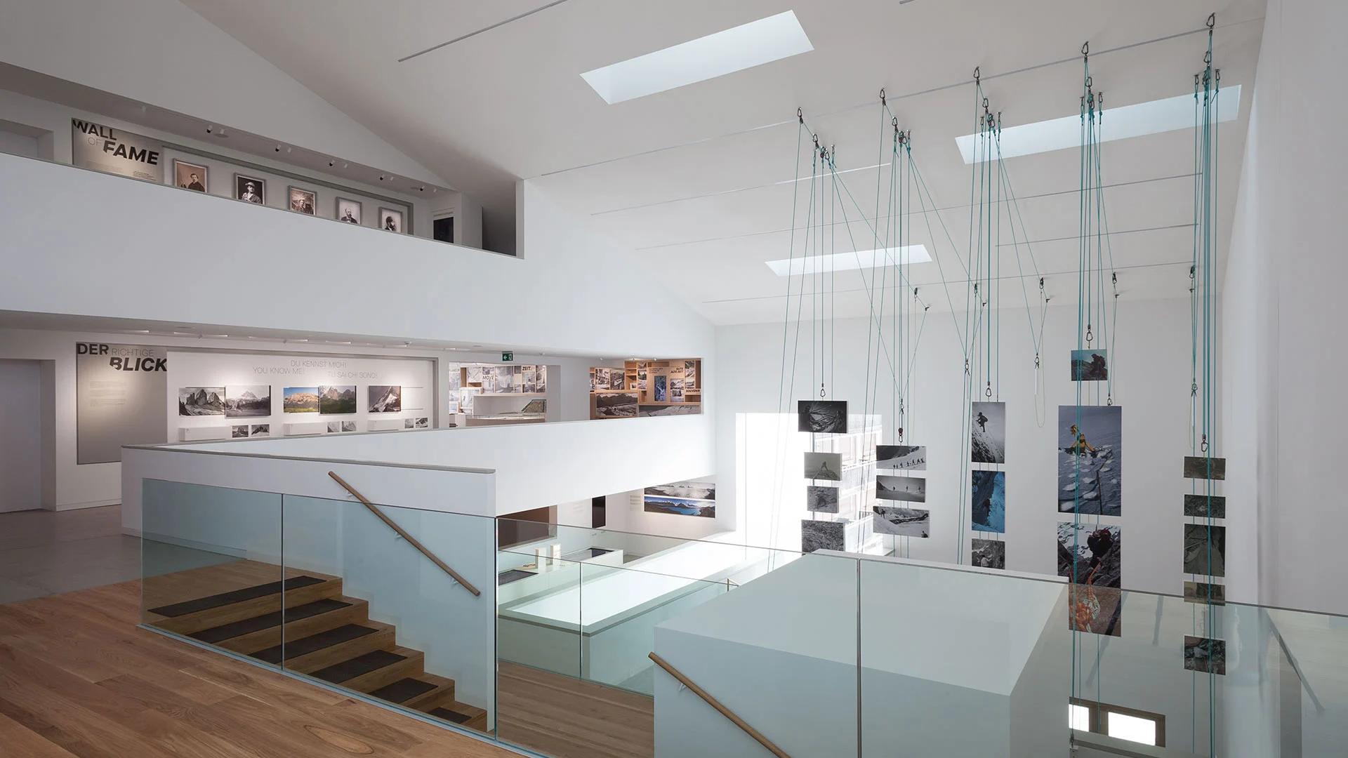 Spazio espositivo luminoso con fotografie sospese a cavi sopra una scala con parapetto in vetro e gallerie lungo le pareti – Lumen Museo della fotografia di montagna, Plan de Corones, Italia, 2018 – architettura