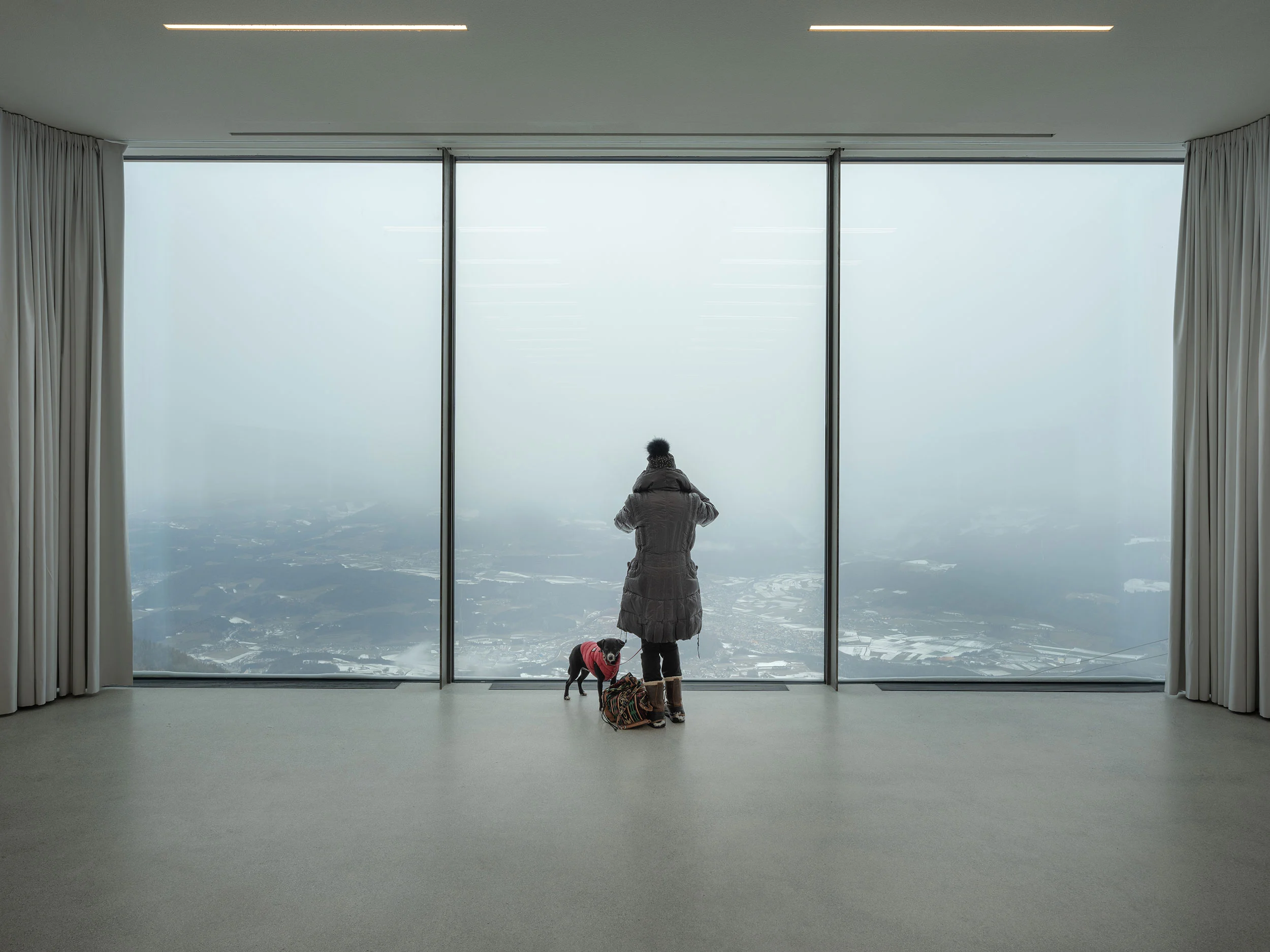 Visitatore con un cane davanti a una vetrata a tutta altezza su un paesaggio montano innevato e nebbioso – Lumen Museo della fotografia di montagna, Plan de Corones, Italia, 2018 – architettura
