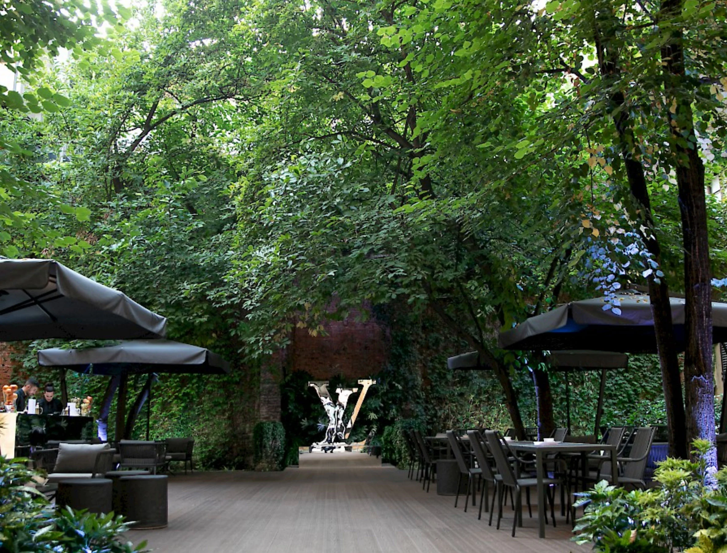 Lounge in giardino sotto alberi alti con ombrelloni grigi, tavoli da esterno e una scultura LV in fondo – Louis Vuitton a Palazzo Clerici, Milano, Italia, 2019 – allestimento espositivo