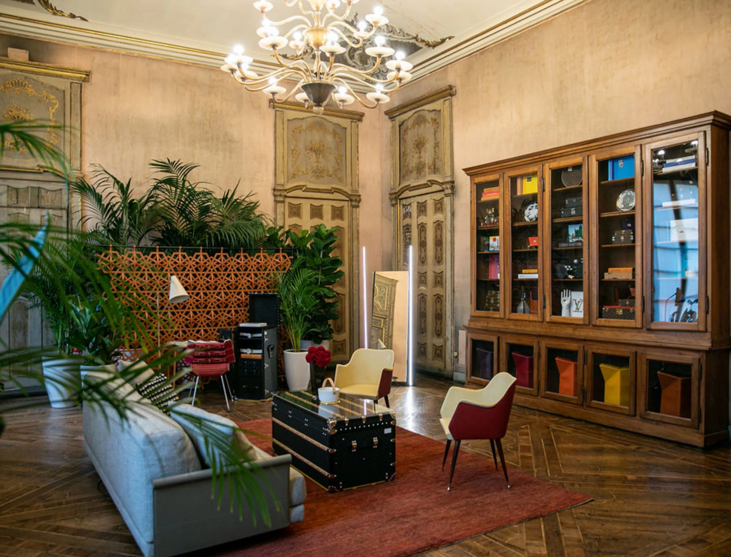 Salone con un lampadario di cristallo, un baule usato come tavolino e una libreria in legno con oggetti – Louis Vuitton a Palazzo Clerici, Milano, Italia, 2019 – allestimento espositivo