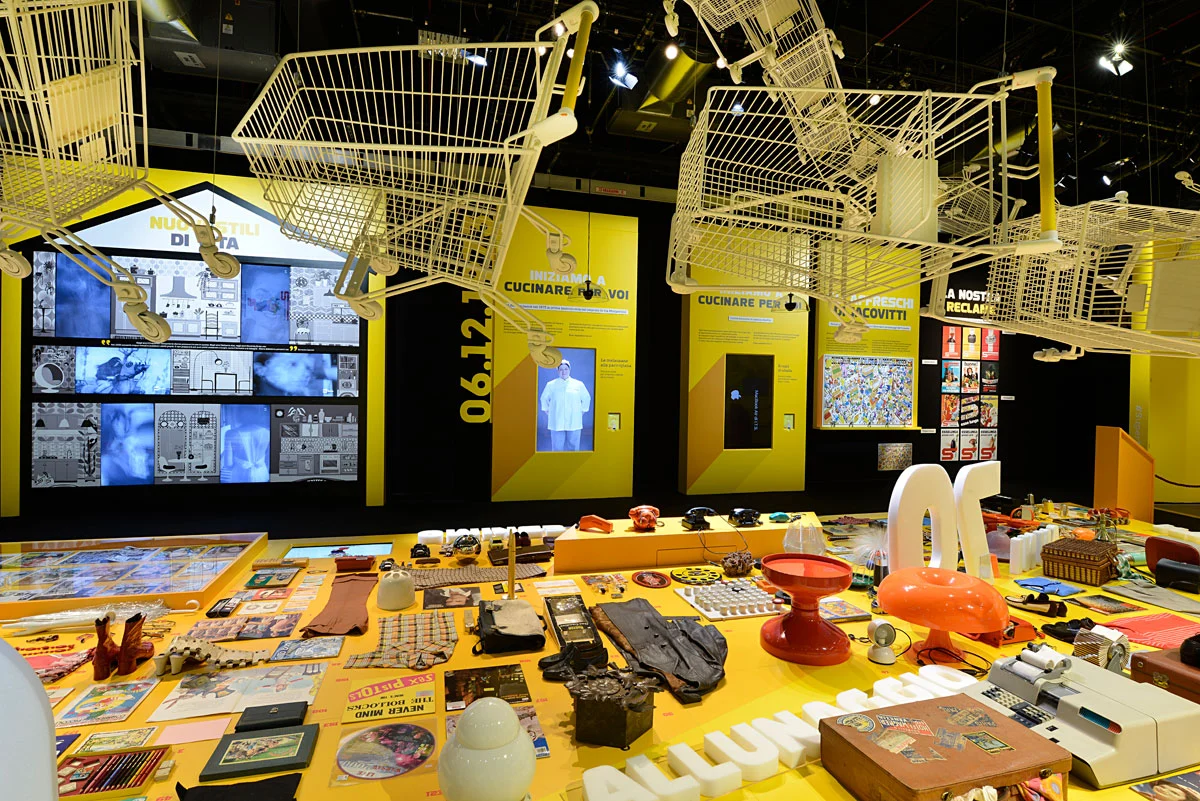 Sala di Supermostra Esselunga con carrelli della spesa appesi al soffitto sopra un tavolo giallo di prodotti d'epoca – Esselunga Supermostra 60° anniversario, Milano, Italia, 2018 – allestimento espositivo