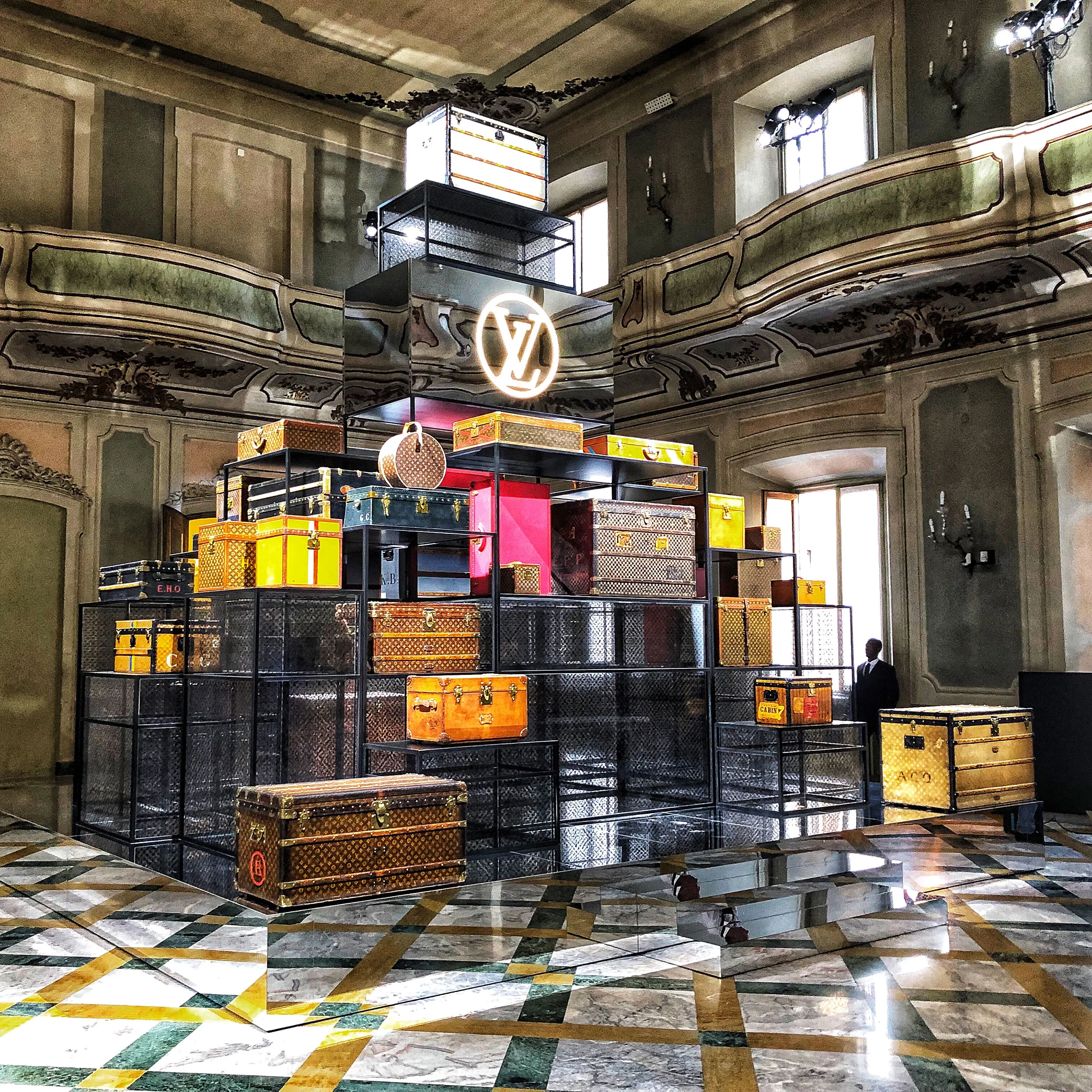 Piramide di bauli Louis Vuitton su scaffali in rete nera sotto un logo LV illuminato, in una sala affrescata – Louis Vuitton a Palazzo Clerici, Milano, Italia, 2019 – allestimento espositivo