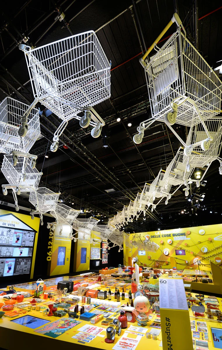Vista verticale di carrelli della spesa bianchi sospesi in una lunga fila sopra tavoli espositivi gialli – Esselunga Supermostra 60° anniversario, Milano, Italia, 2018 – allestimento espositivo