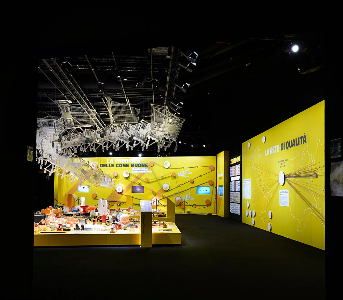 Sala espositiva buia con una nuvola di carrelli sospesi sopra un tavolo giallo e una mappa a parete intitolata La rete di qualità – Esselunga Supermostra 60° anniversario, Milano, Italia, 2018 – allestimento espositivo
