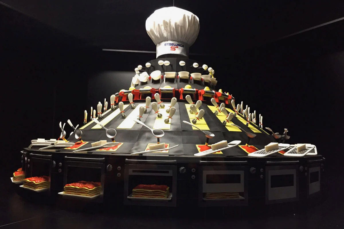 Scultura a gradoni sormontata da un gigantesco cappello da chef Esselunga, con utensili, teglie di lasagne e sportelli di forno – Esselunga Supermostra 60° anniversario, Milano, Italia, 2018 – allestimento espositivo