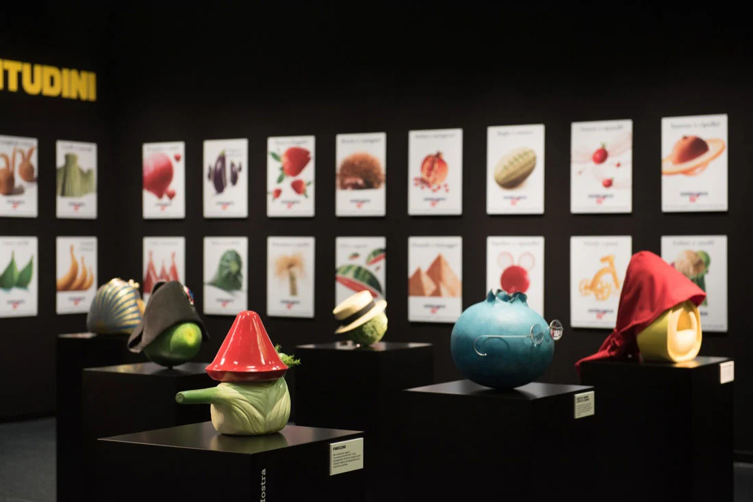 Sculture a forma di ortaggi con cappelli su basamenti neri, davanti a una parete di manifesti illustrati – Esselunga Supermostra 60° anniversario, Milano, Italia, 2018 – allestimento espositivo