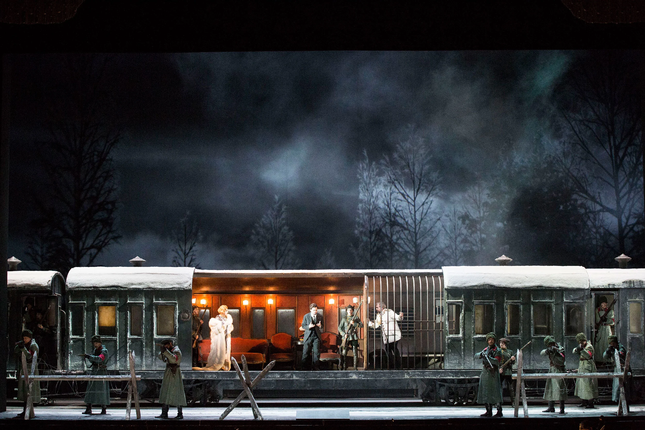 Scenografia di un convoglio ferroviario in un bosco innevato di notte, un vagone salone illuminato con artisti e soldati davanti – Tamerlano al Teatro alla Scala, Milano, Italia, 2017 – scenografia d'opera