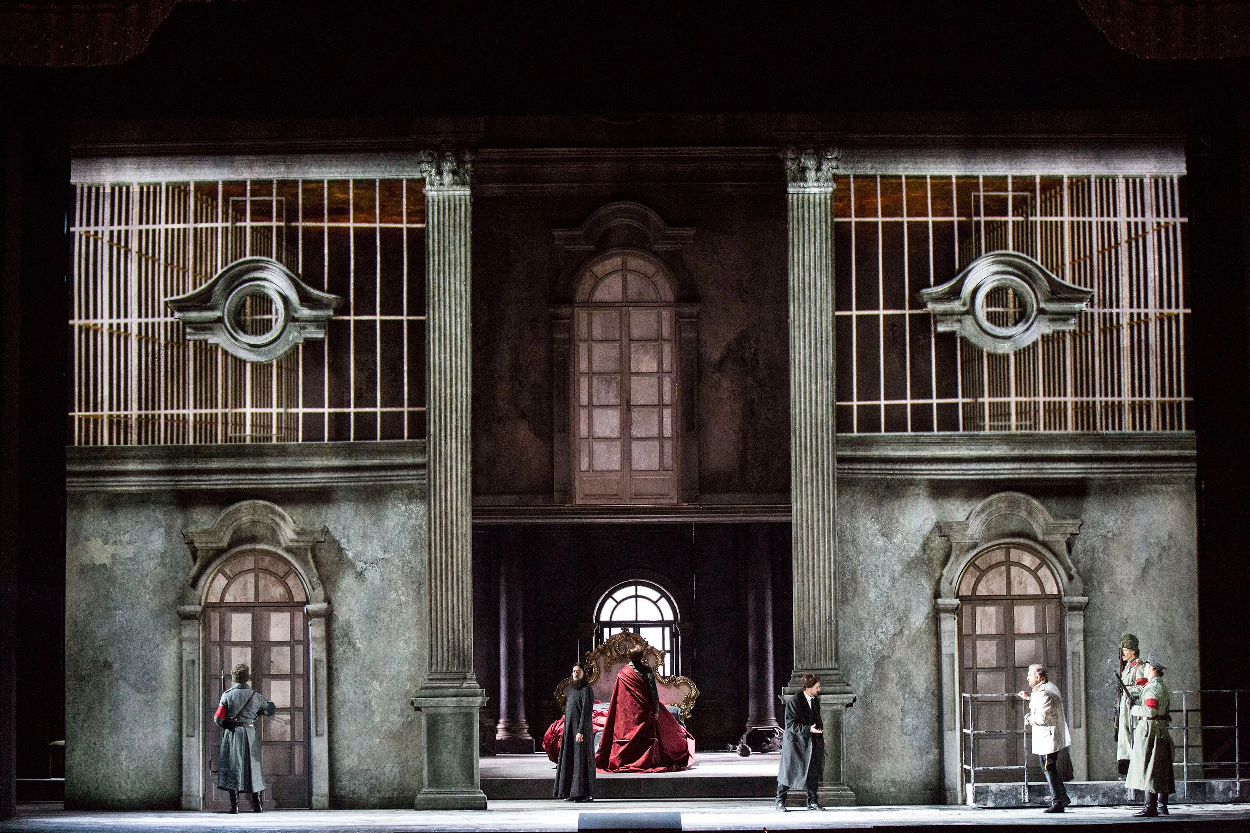 Scenografia di Tamerlano: facciata di palazzo consunta con finestre ad arco e grate, una figura in rosso su un trono al centro – Tamerlano al Teatro alla Scala, Milano, Italia, 2017 – scenografia d'opera