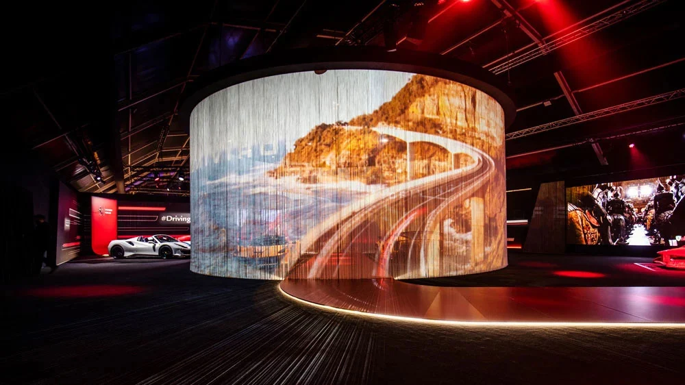 Schermo cilindrico di tende a fili con una strada costiera, circondato da luce in una sala espositiva buia – Universo Ferrari alla Fabbrica del Vapore, Milano, Italia, 2019 – allestimento espositivo
