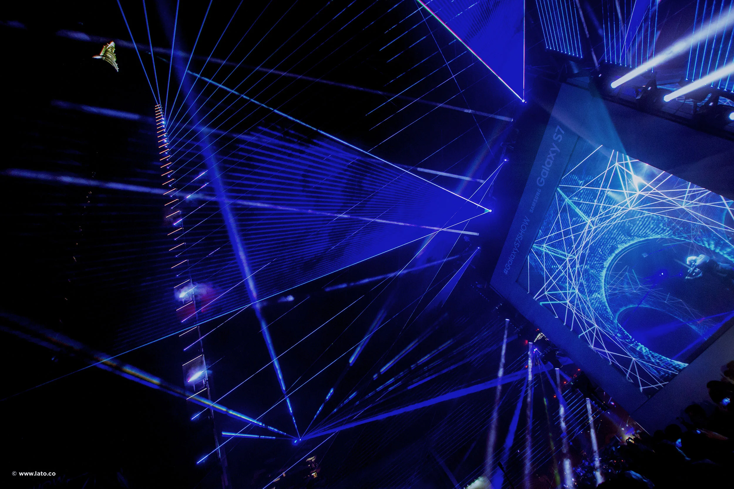 Vista dal basso di linee laser blu che formano disegni geometrici tra gli edifici, accanto a uno schermo video – Samsung Galaxy S7 Laser Show, Milano, Italia, 2016 – progettazione di eventi