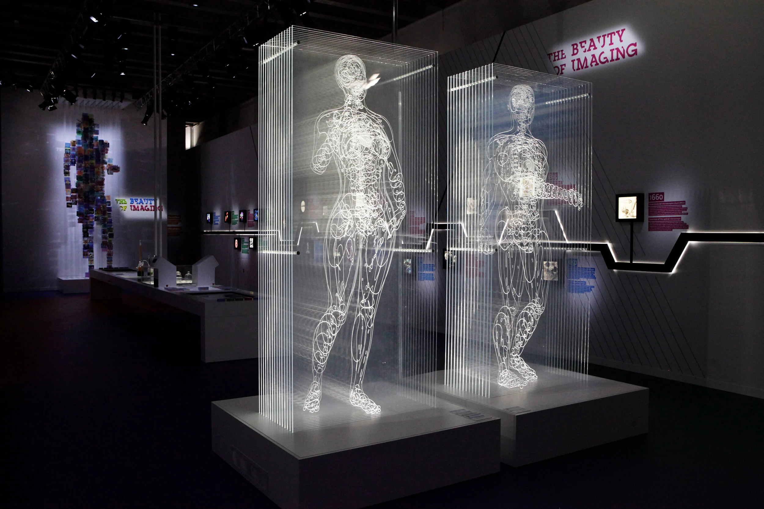 Due figure umane trasparenti incise nella luce su strati verticali, nel buio della mostra The Beauty of Imaging – Bracco The Beauty of Imaging, Milano, Italia, 2017 – allestimento espositivo