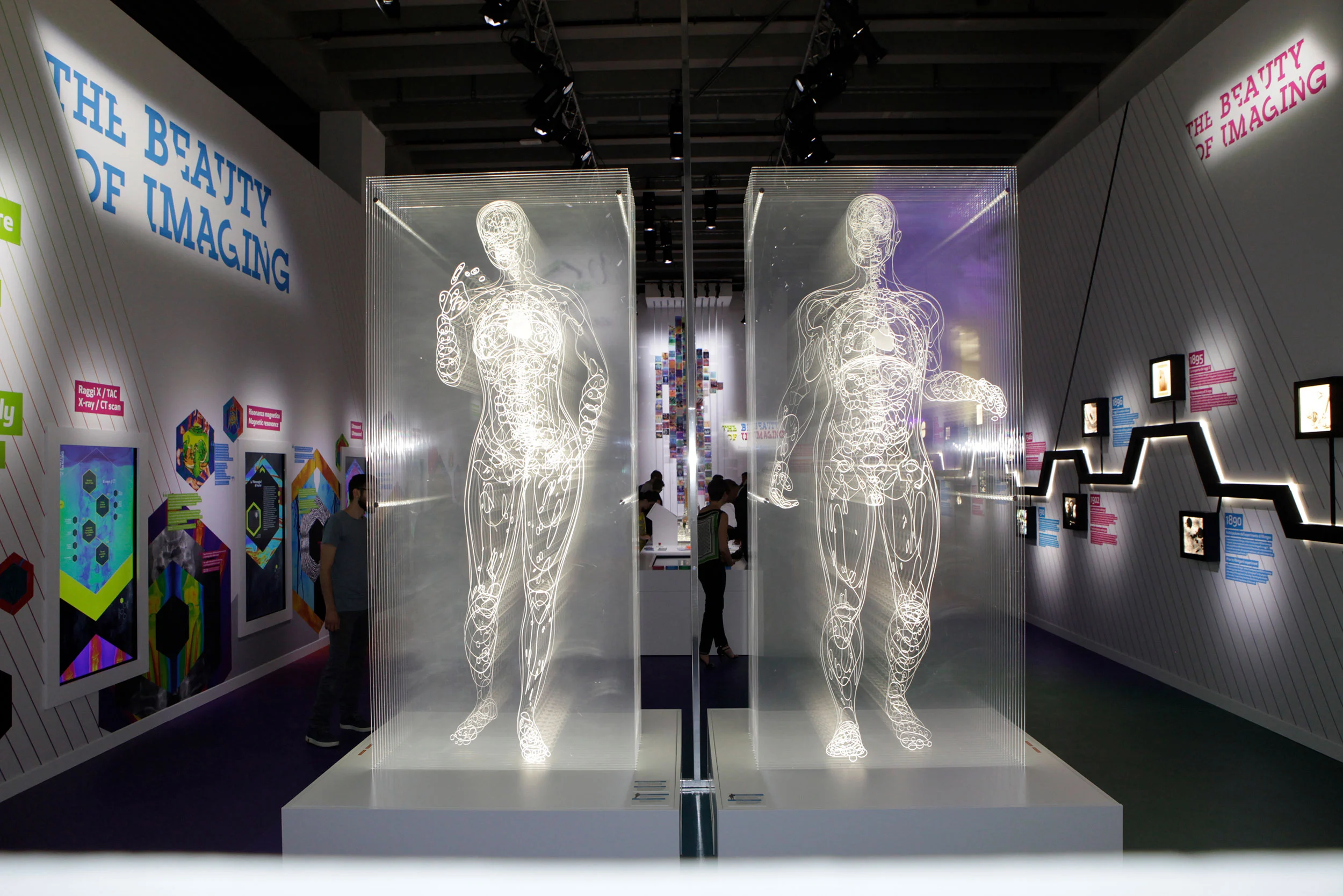 Vista frontale di due figure anatomiche trasparenti luminose, con pannelli colorati e una cronologia lungo le pareti – Bracco The Beauty of Imaging, Milano, Italia, 2017 – allestimento espositivo