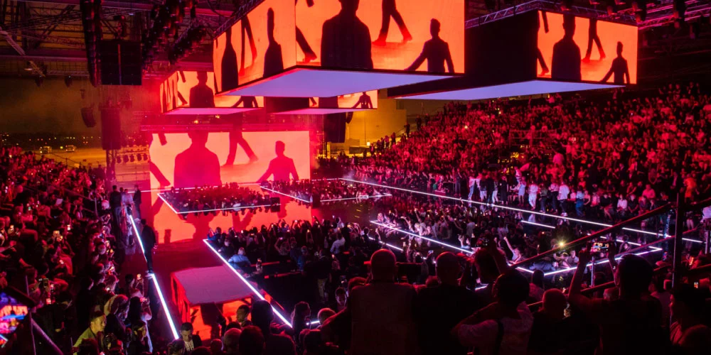 Hangar immerso in luce rosso-arancio, schermi con sagome nere di figure sopra le passerelle – Emporio Armani Boarding SS19, Milano, Italia, 2019 – scenografia per sfilate
