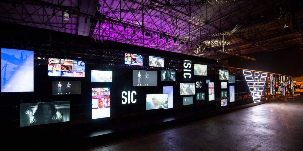 Lunga parete nera di schermi video con le scritte SIC e il logo dell'aquila sotto travature del tetto illuminate di viola – Emporio Armani Boarding SS19, Milano, Italia, 2019 – scenografia per sfilate