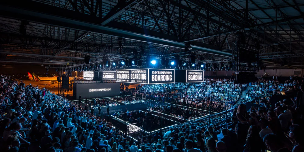 Vista d'insieme dell'hangar con tribune, schermi Emporio Armani Boarding e un aereo sullo sfondo – Emporio Armani Boarding SS19, Milano, Italia, 2019 – scenografia per sfilate