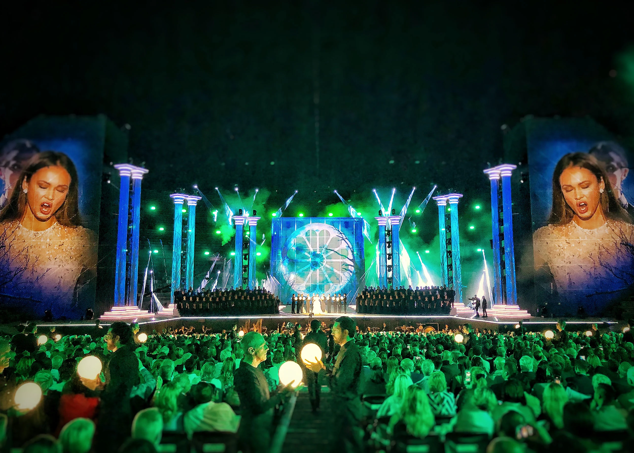 Palco in luce verde con un coro e un pannello a rosone al centro, pubblico con sfere luminose in primo piano – La Notte di Andrea Bocelli all'Arena di Verona, Italia, 2018 – scenografia per concerti
