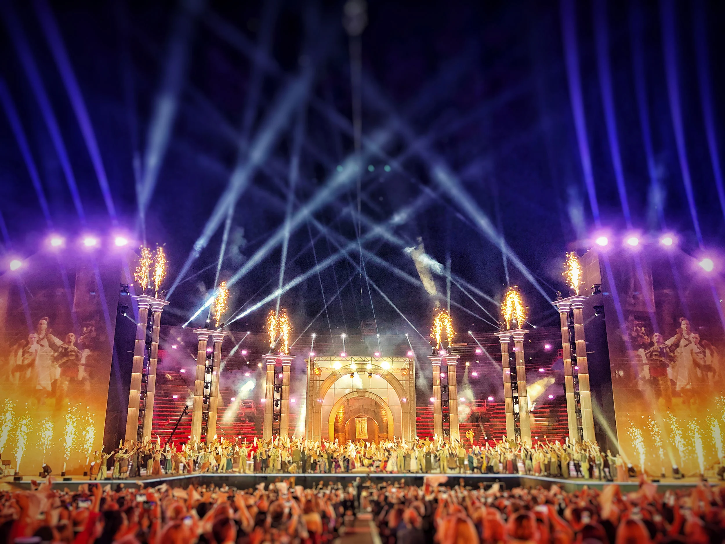 Palco dell'Arena di Verona per La Notte di Andrea Bocelli: colonne con sommità in fiamme, un arco centrale e tutto il cast sotto fasci di luce – La Notte di Andrea Bocelli all'Arena di Verona, Italia, 2018 – scenografia per concerti