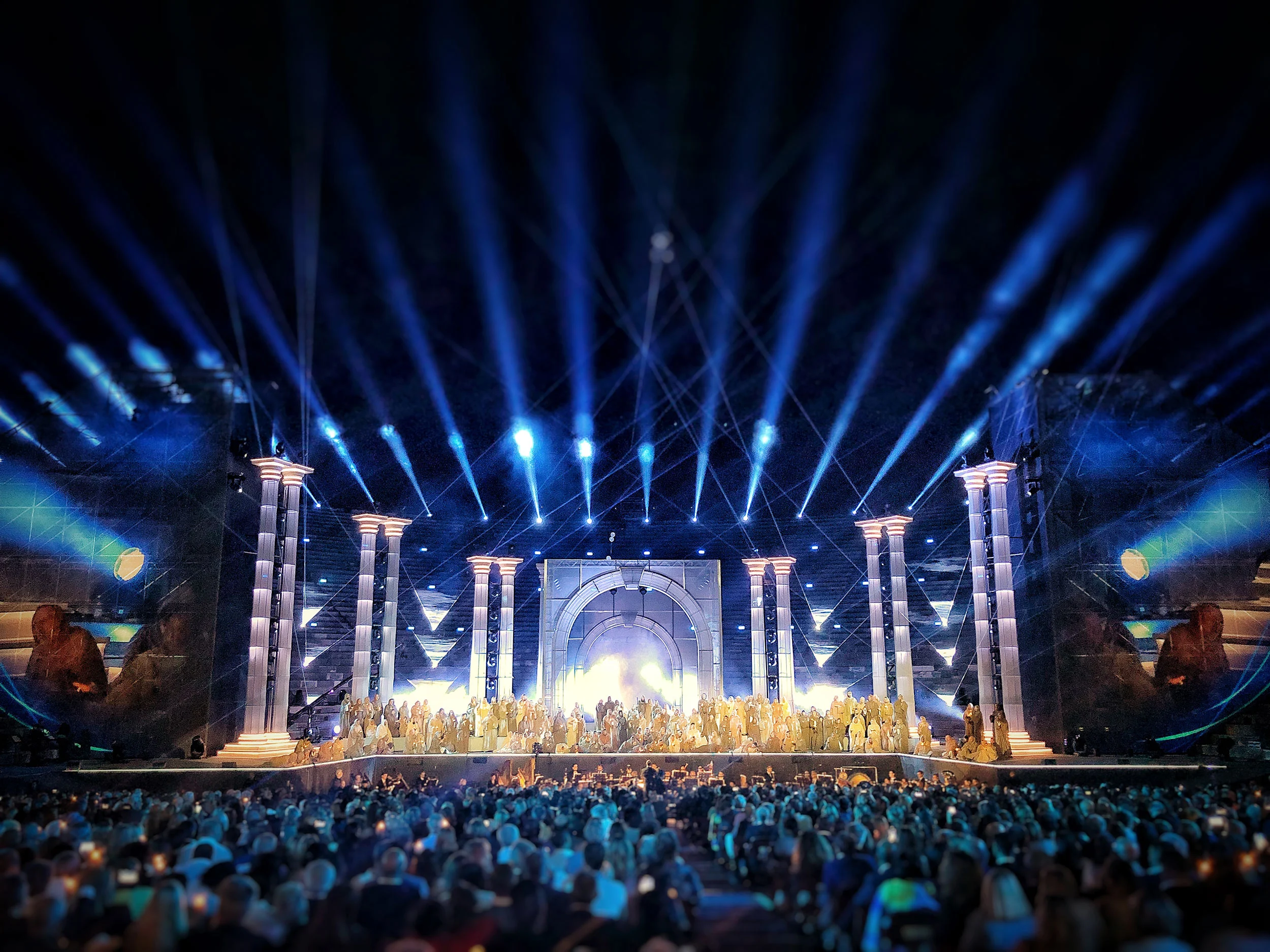 Fasci di luce blu a ventaglio sulla scenografia a colonnato classico, con il cast riunito sotto un luminoso arco centrale – La Notte di Andrea Bocelli all'Arena di Verona, Italia, 2018 – scenografia per concerti