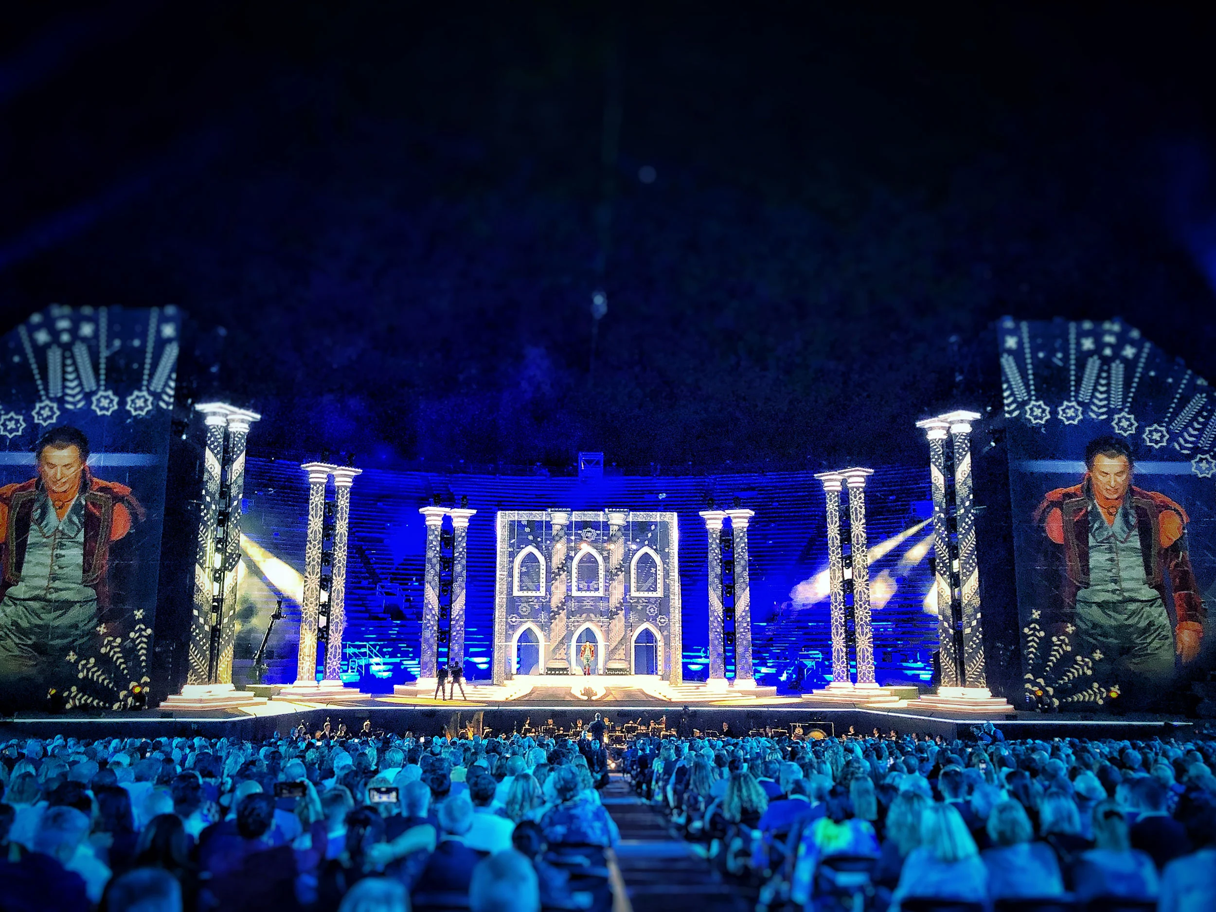 Palco immerso nella luce blu, con colonne decorate, una facciata in stile gotico illuminata e primi piani di un artista sugli schermi laterali – La Notte di Andrea Bocelli all'Arena di Verona, Italia, 2018 – scenografia per concerti