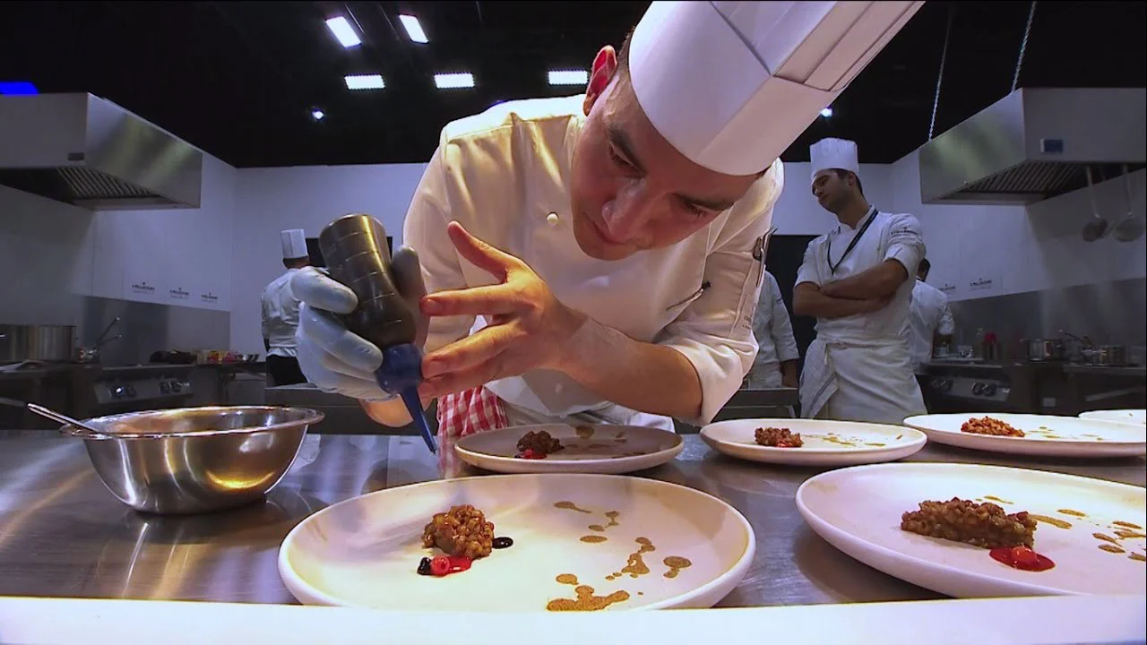 Giovane chef con cappello bianco che impiatta con un biberon da cucina in una cucina professionale – S.Pellegrino Young Chef, Milano, Italia, 2019 – progettazione di eventi