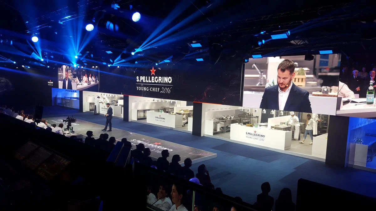 Palco con postazioni di cucina a vista, un grande schermo con un dettaglio e riflettori blu – S.Pellegrino Young Chef, Milano, Italia, 2019 – progettazione di eventi