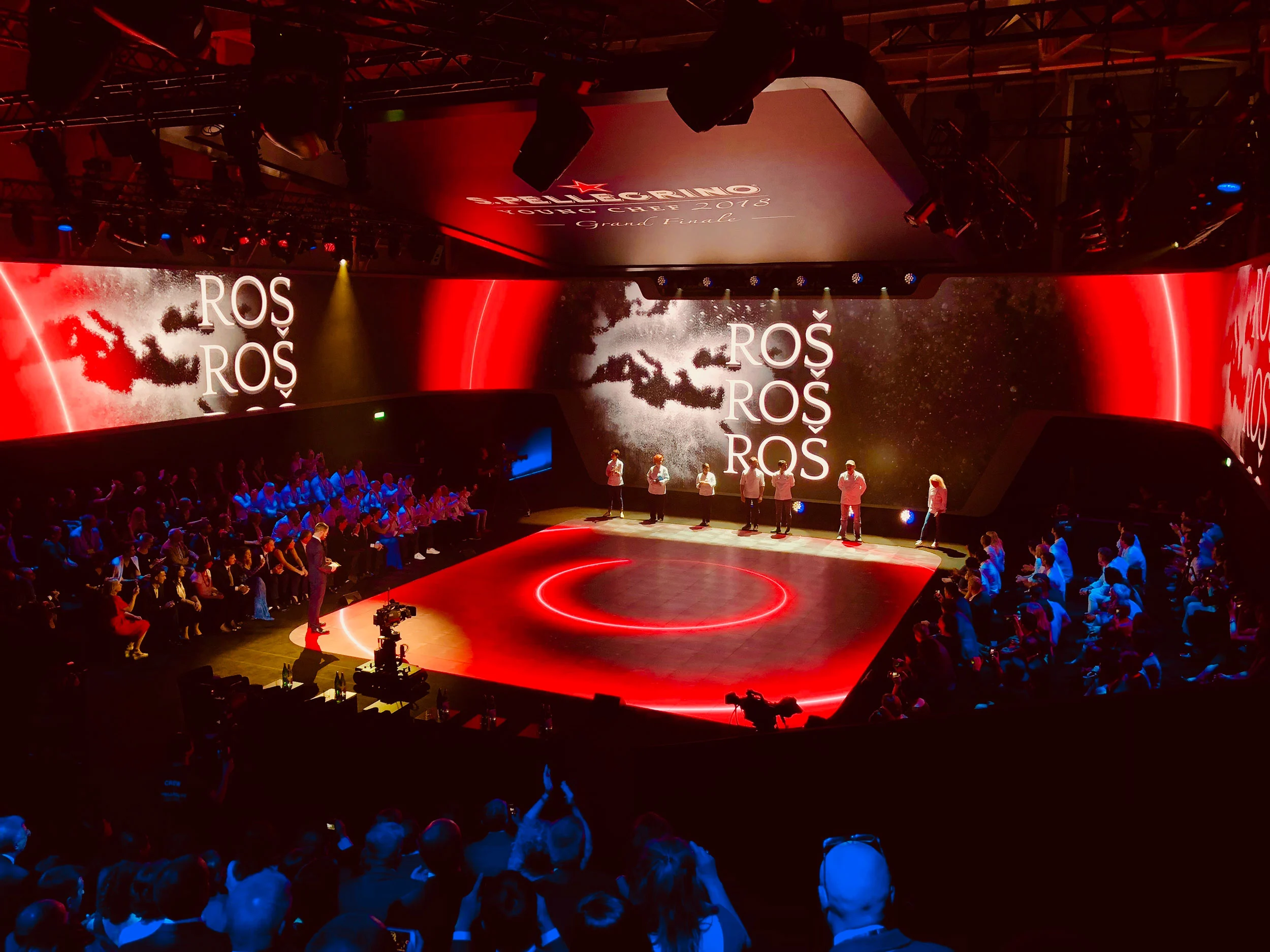 Finale di S.Pellegrino Young Chef: chef su un palco rotondo illuminato di rosso davanti a schermi, pubblico attorno – S.Pellegrino Young Chef, Milano, Italia, 2019 – progettazione di eventi