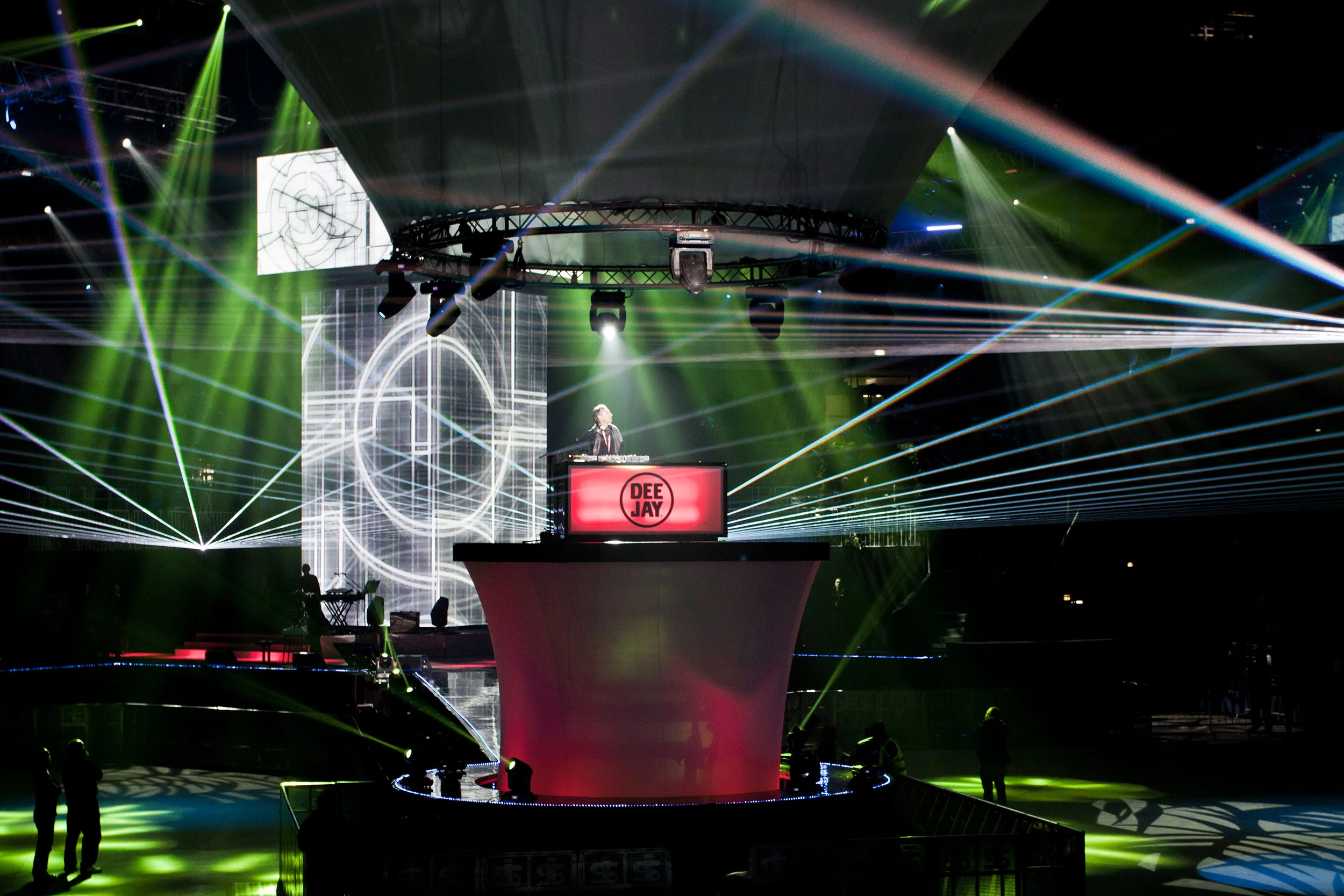 Consolle del DJ con l'insegna Deejay su un podio rotondo rosso, attraversata da fasci laser verdi – Festa per i 30 anni di Radio Deejay, Assago, Italia, 2019 – scenografia per concerti