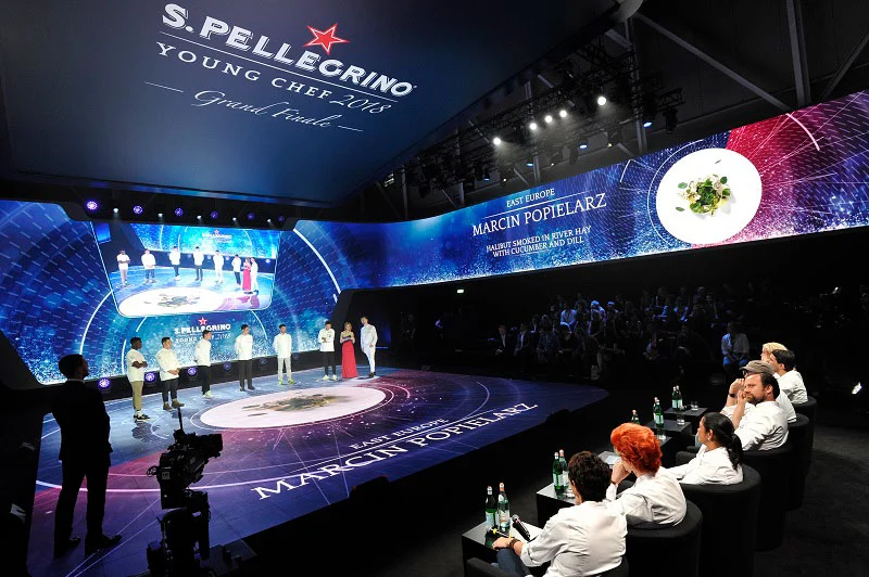 Palco della finale con una parete LED curva che mostra un piatto e i nomi dei finalisti, la giuria seduta a destra – S.Pellegrino Young Chef, Milano, Italia, 2019 – progettazione di eventi
