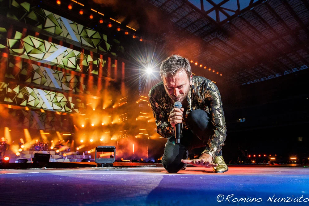 Cesare Cremonini in ginocchio con un microfono, alle spalle schermi video geometrici in luce arancione – Cesare Cremonini Stadi 2018, Italia – scenografia per concerti
