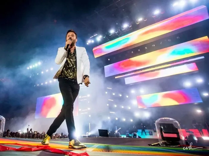 Cesare Cremonini canta sul bordo del palco in giacca bianca, dietro di lui barre video colorate – Cesare Cremonini Stadi 2018, Italia – scenografia per concerti