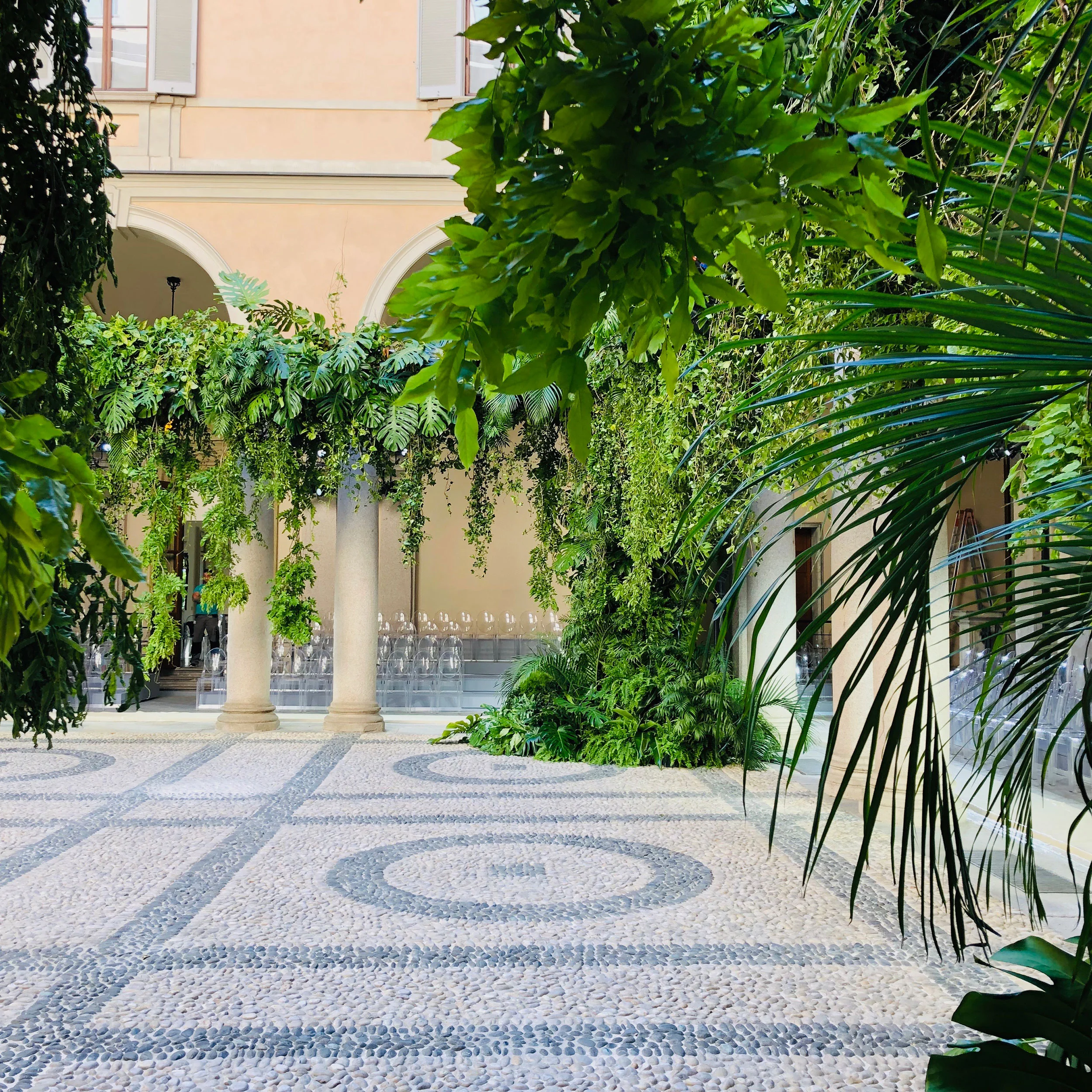 Chiostro di Palazzo Orsini con pavimento in ciottoli, colonne coperte di piante tropicali e file di sedie trasparenti – Giorgio Armani SS20 Uomo e Donna, Milano, Italia, 2019 – scenografia per sfilate