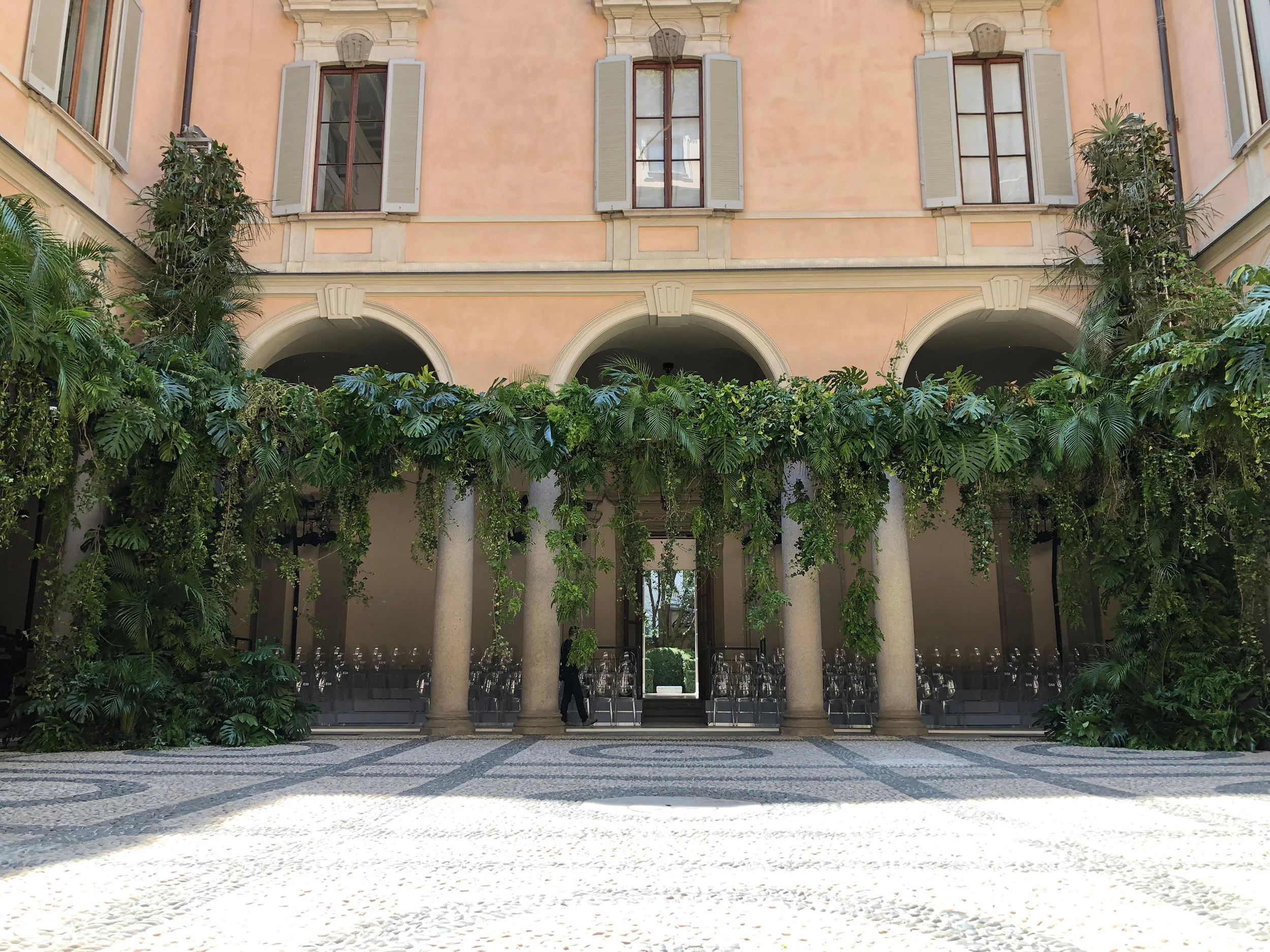 Cortile di Palazzo Orsini con piante tropicali che ricadono dal colonnato e pavimento in ciottoli – Giorgio Armani SS20 Uomo e Donna, Milano, Italia, 2019 – scenografia per sfilate