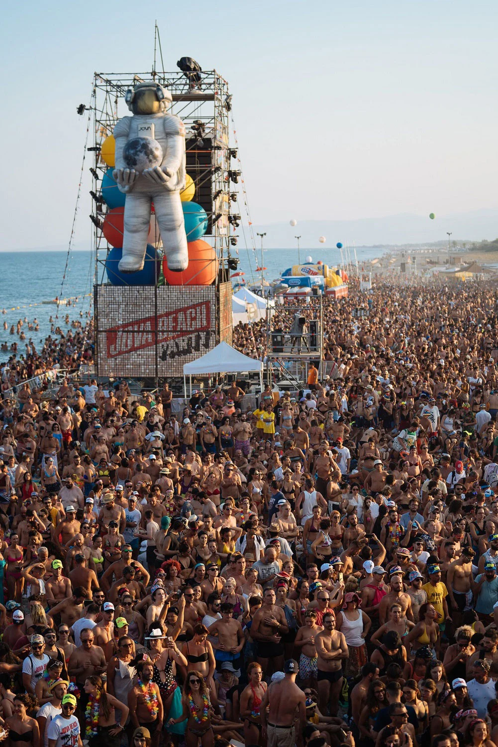 Enorme astronauta gonfiabile su una torre di tubi sopra un pubblico che si estende lungo la spiaggia fino al mare – Jova Beach Party 2019, Italia – scenografia per concerti